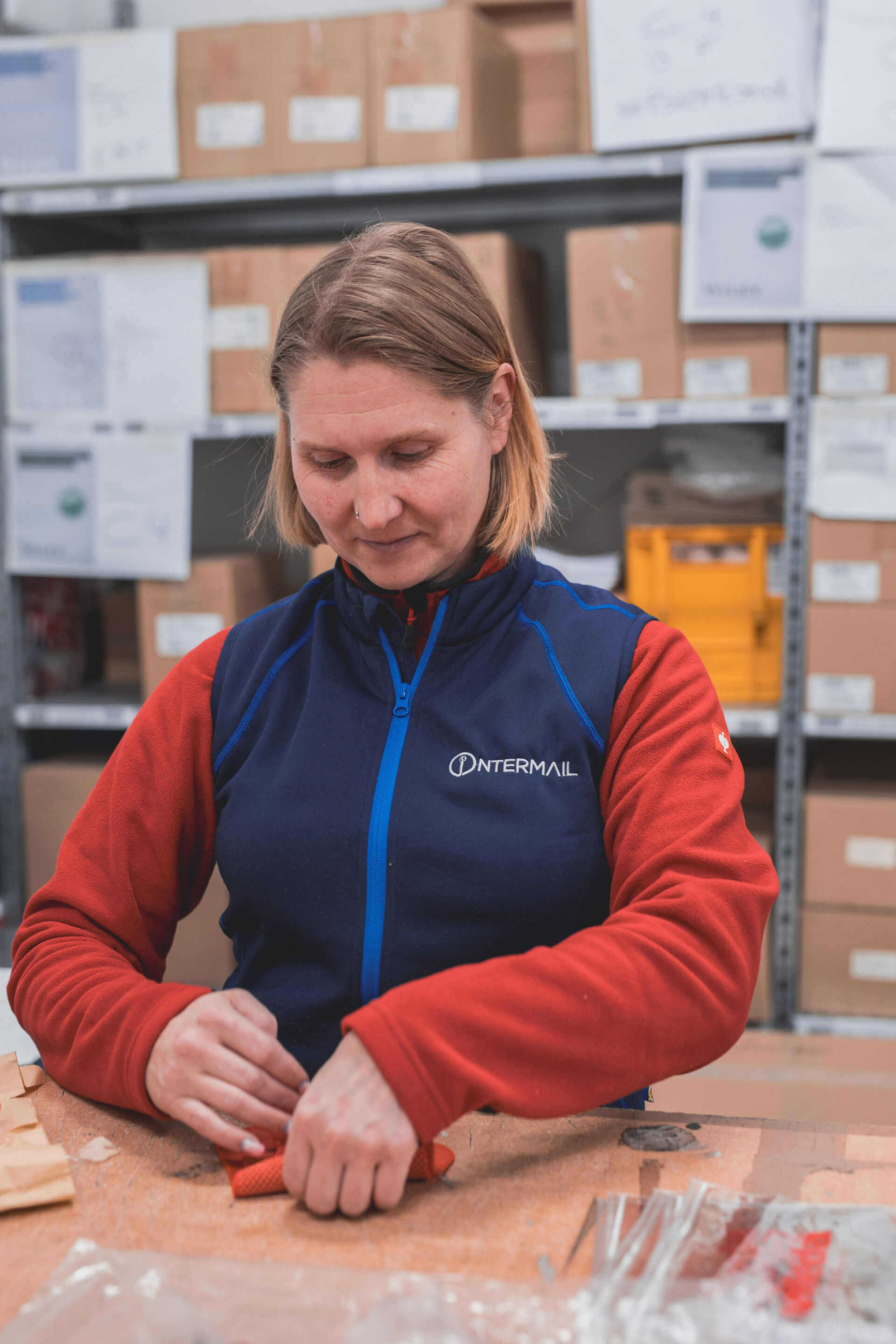 Frau in blauer Intermail-Weste und rotem Shirt arbeitet konzentriert an einem Tisch im Lager.