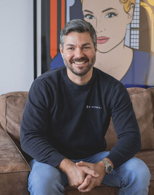 Lächelnder Mann mit Bart und Intermail-Pullover sitzt auf einem braunen Sofa vor einem Pop-Art-Bild.