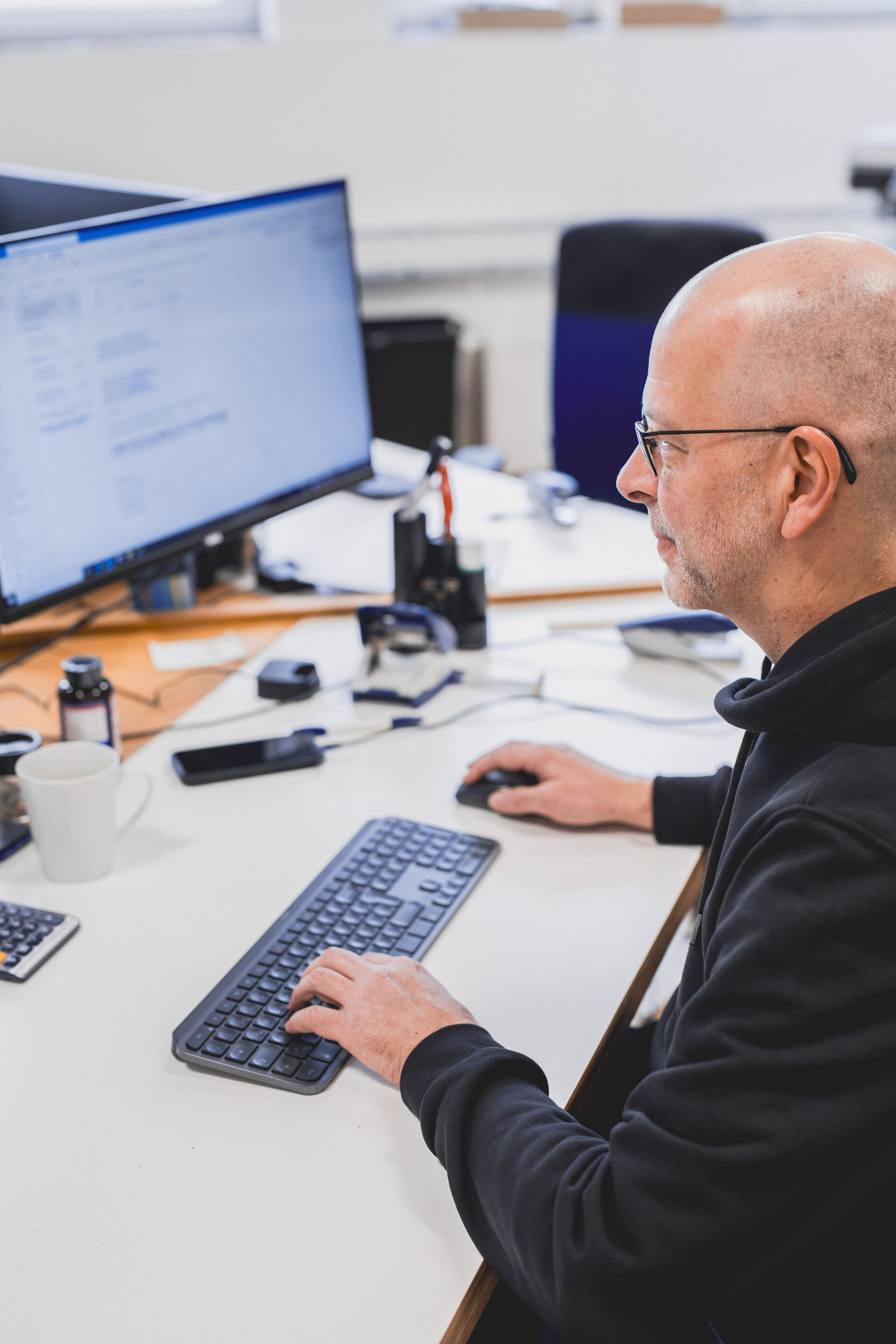 Glatzköpfiger Mann mit Brille in einem dunklen Hoodie arbeitet konzentriert an seinem Computer am Schreibtisch.