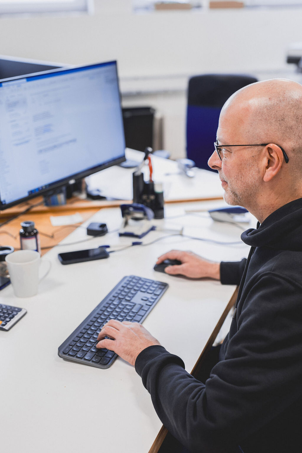 Glatzköpfiger Mann mit Brille in einem dunklen Hoodie arbeitet konzentriert an seinem Computer am Schreibtisch.