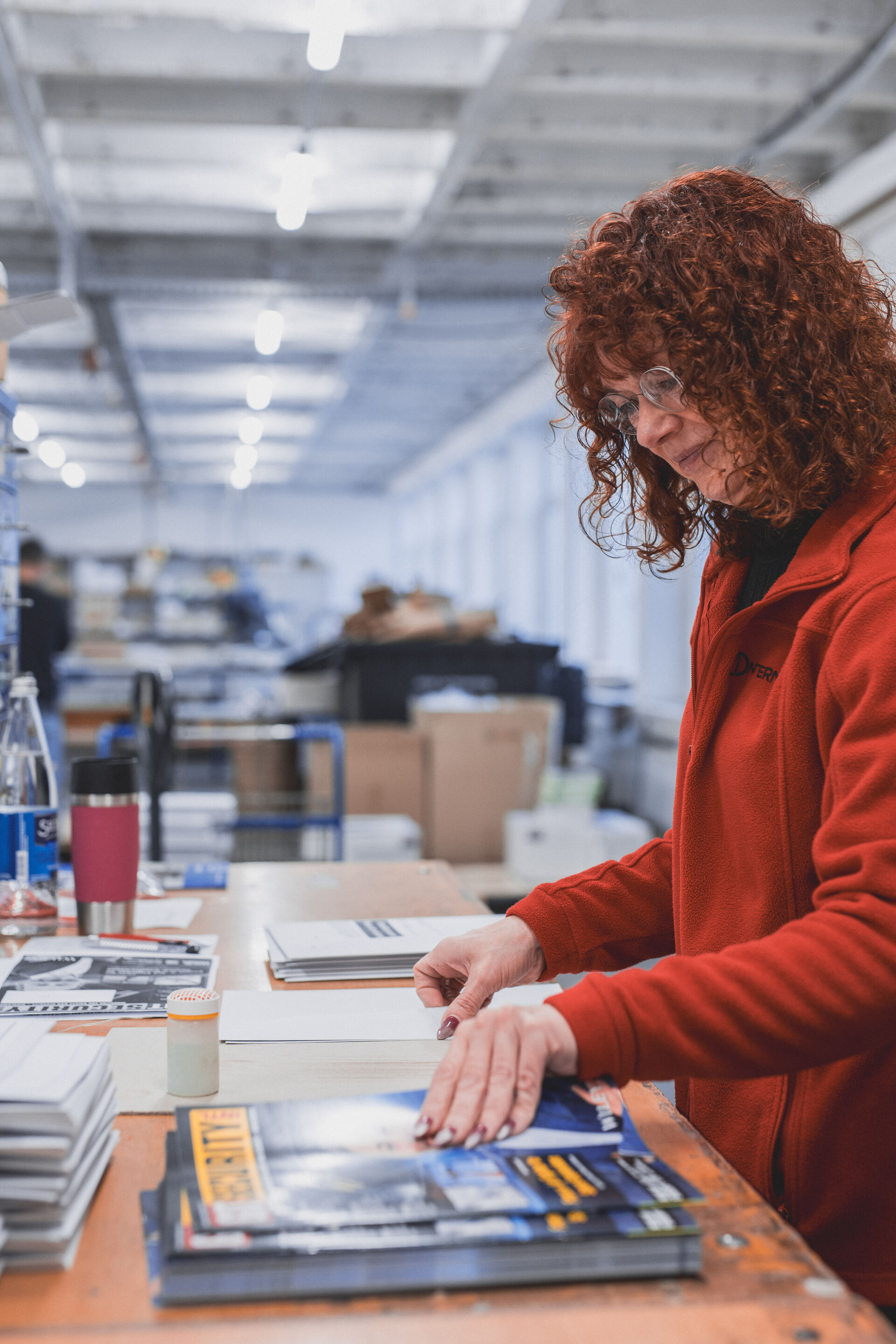 Frau mit roten Locken und Brille in roter Jacke sortiert an einem Tisch im Lager Zeitschriften.
