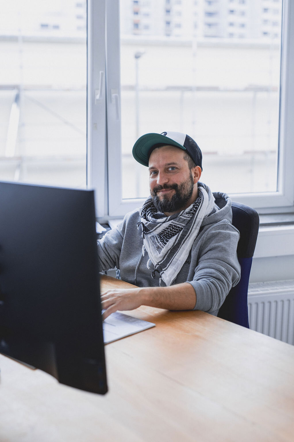 Bärtiger Mann mit Kappe und Schal sitzt lächelnd an seinem Schreibtisch im Büro vor einem Monitor.