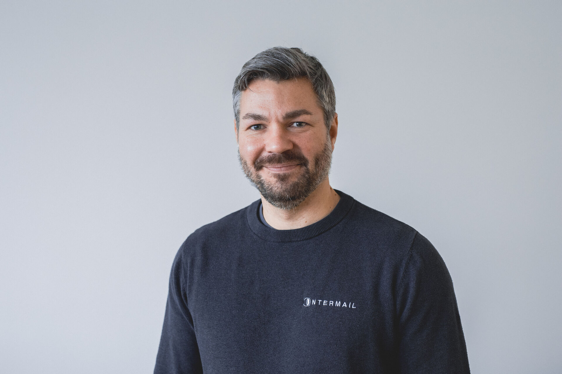 Lächelnder Mann mit grauem Haar, Bart und dunklem Intermail-Pullover.