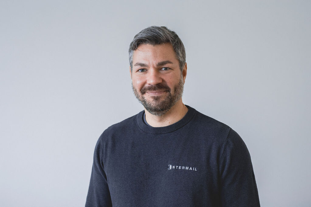 Lächelnder Mann mit grauem Haar, Bart und dunklem Intermail-Pullover.